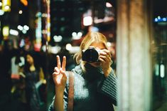 a woman taking a selfie with her camera in the city at night, holding up two fingers