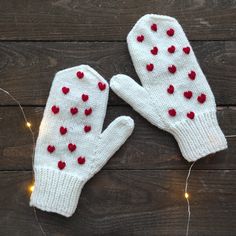 two white mittens with red hearts on them sitting on a wooden floor next to string lights