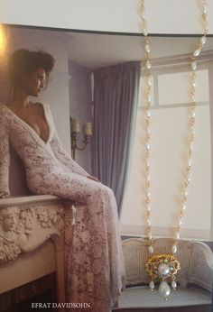 a woman sitting on top of a bed next to a window with a necklace hanging from it's side