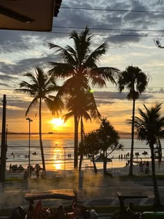 the sun is setting behind palm trees and people are on the beach in the distance