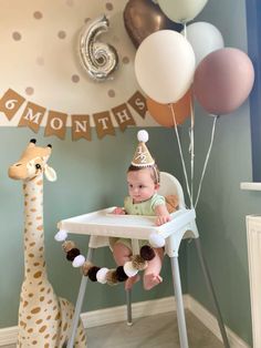 a baby sitting in a high chair next to a giraffe and balloon garland