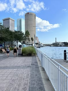 two people walking down a sidewalk next to the water with tall buildings in the background