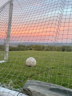 two soccer balls sitting in the grass behind a net on a grassy field at sunset