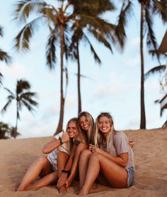 three women sitting on the sand with palm trees in the backgroung behind them