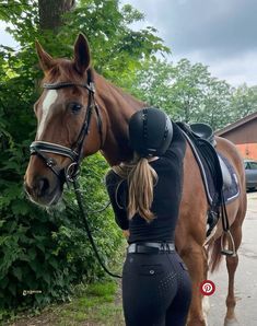 a woman in black riding gear standing next to a brown horse on a road with trees and bushes behind her