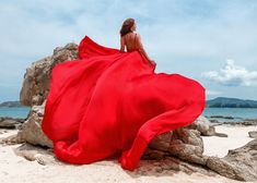 a woman in a long red dress sitting on rocks at the beach looking out to sea