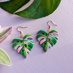 two green and pink leaf shaped earrings sitting on top of a white table next to a plant