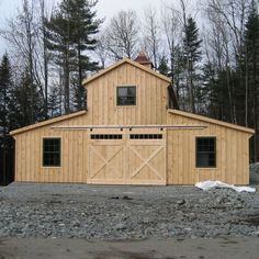 a large wooden barn sitting on top of a gravel covered field next to trees and rocks