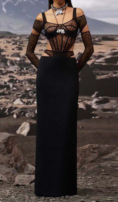 a woman in a long black dress standing on top of a rocky hill with her hands on her hips