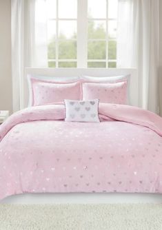 a bed with pink comforter and pillows in a white room next to a window