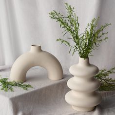 two white vases sitting on top of a table covered in greenery next to each other