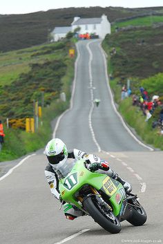 a man riding a green motorcycle down a curvy road next to a lush green hillside