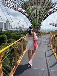 a woman in white dress walking across a bridge with trees and ferris wheel in the background