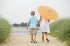 two people walking on the beach with an umbrella over their heads and sand dunes behind them