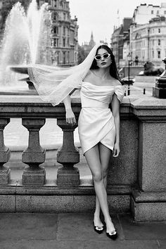 black and white photo of woman in dress with veil over head leaning on railing near fountain