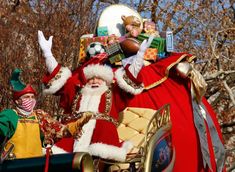 two people dressed as santa clause riding in a horse drawn carriage with presents on it