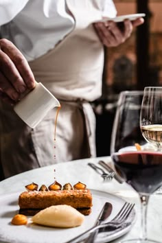 a person pouring sauce on some food on a plate with wine glasses and utensils