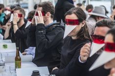 many people are sitting at tables with their faces covered by paper and blindfolds