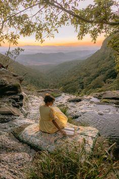 a woman sitting on top of a blanket next to a river in the mountains at sunset