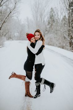 two women hugging each other in the middle of snow covered road with trees behind them