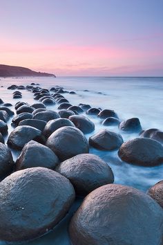 rocks lined up on the beach in front of the ocean at sunset, with pink and blue sky