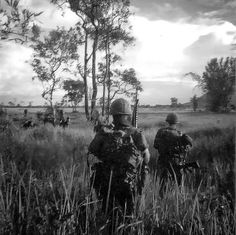 black and white photograph of two soldiers walking through tall grass with trees in the background