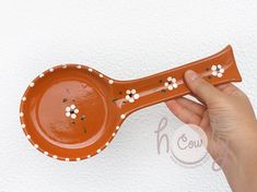 a hand holding an orange ceramic spoon with white flowers on the handle and holes in it