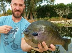 a man holding up a large fish in his hand while standing next to a pond