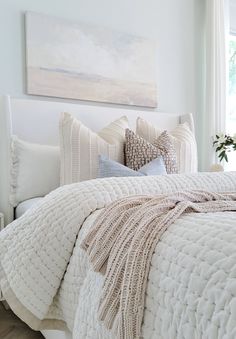 a white bed with pillows and blankets in a bedroom next to a painting on the wall