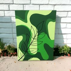 a painting of a green leaf on a white brick wall in front of some plants