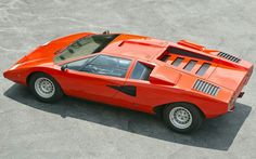 an orange sports car parked in a parking lot next to a person's hand