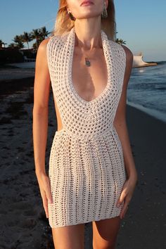 a woman standing on top of a sandy beach next to the ocean wearing a white crocheted dress