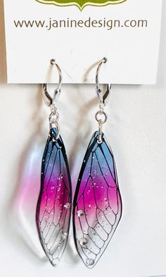 a pair of butterfly wings are hanging from the ear hooks on a white background with a business card behind them