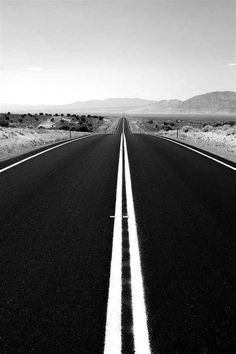 a black and white photo of an empty road in the desert with mountains in the background