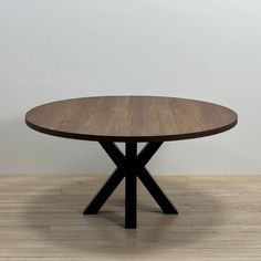 a round wooden table with black cross legs on a hard wood flooring area in front of a white wall