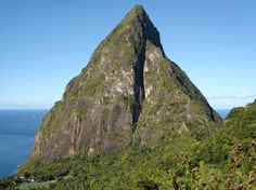 a tall mountain towering over a lush green forest covered hillside next to the ocean on a sunny day