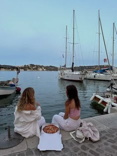 two women are sitting on the dock eating pizza and drinking wine in front of sailboats