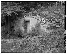 black and white photograph of an old stone house in the woods with ivy growing around it