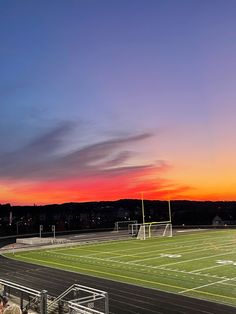 an empty football field at sunset with the sun setting in the distance and people sitting on the bleachers