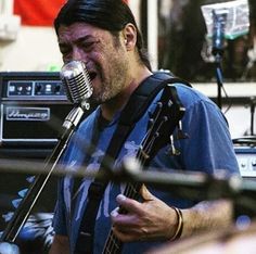 a man singing into a microphone while holding a guitar in his right hand and wearing a blue t - shirt