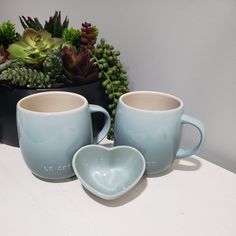 two mugs sitting next to each other on a table with succulents in the background