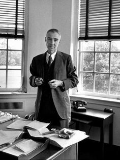 black and white photograph of man standing in front of desk with papers on it, looking at camera