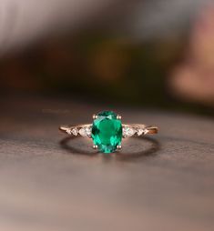 an oval cut emerald ring with three diamonds on the side, sitting on top of a wooden table