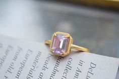 This ring features a central rectangular gemstone with a soft pink hue, cut in an emerald style with step facets that create a classic and elegant look. The stone is set in a gold band with four prongs securing it at each corner, and the setting is bordered by a decorative, textured frame that adds to the ring's vintage appeal. The overall design combines simplicity and sophistication, making it a charming and timeless piece. Details Isabella Rectangular Crystal Vintage Ring Adjustable opening Gold band Rare, antique design Reyis Jewelry Isabella Collection Elegant Ring With Faceted Rectangular Stone, Gold Rectangular Crystal Ring, Elegant Crystal Ring With Rectangular Stone, Elegant Crystal Ring With Rectangular Gemstone, Elegant Gold Rectangular Crystal Ring, Gold Rectangular Amethyst Ring For Wedding, Gold Amethyst Rectangular Ring For Wedding, Rectangular Gold Amethyst Wedding Ring, Formal Pink Rings With Rectangular Stone