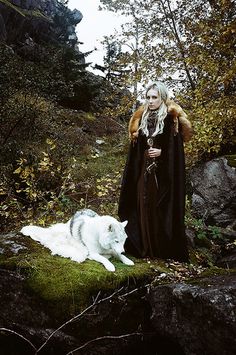 a woman in a long black cloak standing next to a white cat on a rock