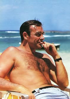 a shirtless man sitting in a chair on the beach talking on his cell phone