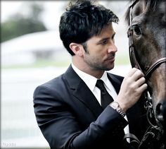 a man in a suit and tie standing next to a horse with his hand on the bridle