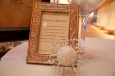 a table topped with a wine glass filled with white flowers and a framed card on top of it