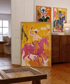 three paintings on the wall in a room with wooden floors and white walls, along with a coffee table