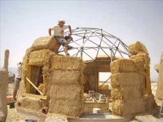 a man standing on top of a pile of hay next to a building made out of straw bales